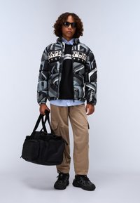 Doudoune avec un motif géométrique gris et noir, dot�ée de logos ; associée à un sweatshirt noir, un pantalon cargo beige et un sac de sport noir.