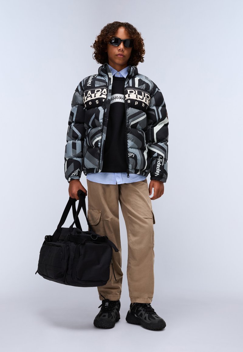 Doudoune avec un motif géométrique gris et noir, dotée de logos ; associée à un sweatshirt noir, un pantalon cargo beige et un sac de sport noir.