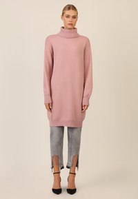 Rosa gestricktes Sweaterkleid mit hohem Rollkragen, lockerer Passform und langen Ärmeln, kombiniert mit grauen cropped Jeans mit Fransen und Verzierungen.
