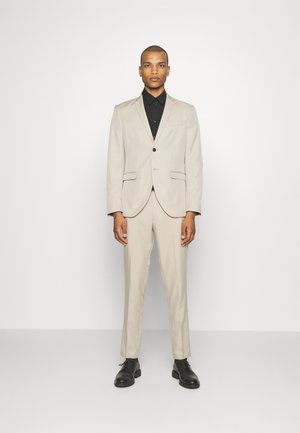 Abito beige con giacca a risvolto e pantaloni coordinati. Camicia nera con bottoni sotto, abbinata a scarpe eleganti nere. Tessuto morbido.