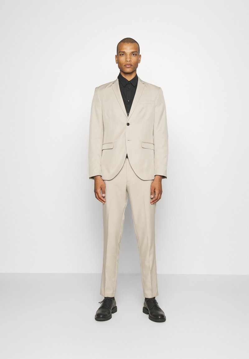 Abito beige con giacca a risvolto e pantaloni coordinati. Camicia nera con bottoni sotto, abbinata a scarpe eleganti nere. Tessuto morbido.