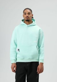 Mintgroene hoodie van zachte stof met een voorzak, voorzien van een klein logo op de borst en een capuchon met trekkoord. Gecombineerd met een donkere broek.