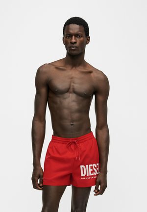 Homme musclé debout de face portant un short de bain rouge Diesel avec logo blanc, sur un fond clair uni.