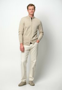 Cardigan beige con cremallera y cuello alto, combinado con pantalones claros y zapatillas beige, exhibido en un modelo frente a un fondo liso.
