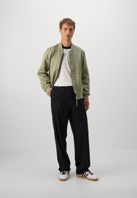 Homme portant une veste bomber verte claire sur un t-shirt à côtes blanc, un pantalon noir ample et des baskets blanches avec des rayures noires, se tenant avec une main dans la poche.