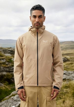 Mees, kes kannab beeži välisjopet ja taskupükse, seisab kivises, rohelises maastikus pilvise taeva all.