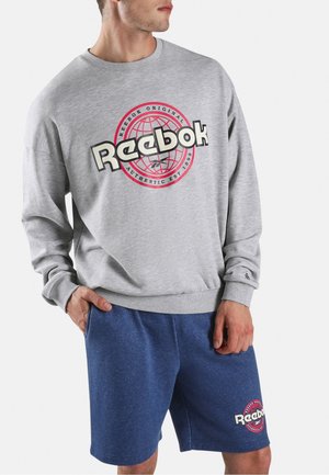 Mann trägt grauen Reebok-Sweatshirt mit rotem Logo und blauem Reebok-Shorts, steht mit einer Hand in der Tasche vor einfarbigem Hintergrund.