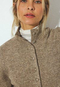 Gilet en tricot marron avec un col haut, doté d'une fermeture à boutons et d'une texture douce. Porté sur un col roulé blanc.
