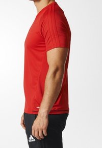 Hombre con una camiseta deportiva roja de manga corta y pantalones negros de Adidas de pie de lado contra un fondo blanco.