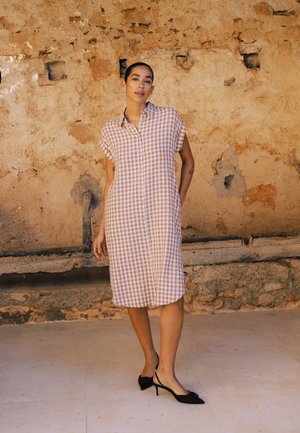 Kobieta z zaczesanymi do tyłu włosami, ubrana w bezrękawową, zapinaną na guziki sukienkę w beżowo-białą kratę oraz czarne czółenka na niskim obcasie, stojąca na tle zniszczonej ściany.