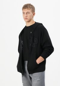 Young man wearing black zip-up hoodie with pocket and grey ripped jeans, standing with one hand in pocket against white background.
