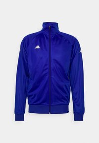 Blå sportjacka av slätt, syntetiskt material. Har hög krage, långa ärmar, dragkedja och vita logodetaljer på bröstet och ärmarna.