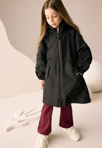 Veste légère noire avec fermeture éclair à l'avant, taille élastique et capuche. Associée à un pantalon de survêtement bordeaux et des baskets blanches.