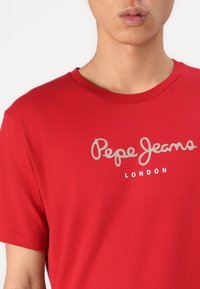 Camiseta de algodón roja con cuello redondeado y mangas cortas. Logo "Pepe Jeans" en gris claro encima de "LONDON" en blanco.