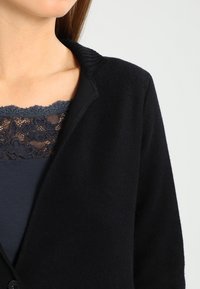 Musta neuletakki, jossa on resorikaulus, pehmeä rakenne ja napillinen suljenta. Alla näkyy tummansininen yläosa, jossa on pitsiyksityiskohtia.