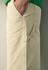 Pantalons beiges en coton avec une poche latérale, dotés de coutures décoratives et d'une texture lisse. Présentés sur un fond neutre.