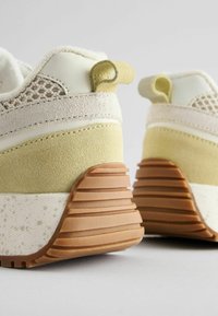 Les baskets présentent un dessus en suède crème et vert pâle avec des accents en mesh. La semelle en caoutchouc texturé a des rainures horizontales et une semelle intermédiaire tachetée.