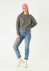 Grijze gebreide trui, blauwe jeans met een iets cropped pasvorm, en witte enkelboots met een gestructureerde zool, afgewerkt met een roze geribde beanie.