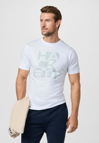 Hombre con camiseta blanca gráfica y pantalones azul marino, sosteniendo una sudadera beige doblada, mirando hacia un lado contra un fondo liso.