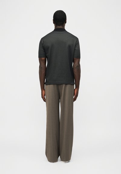 Άνδρας που στέκεται με την πλάτη γυρισμένη, φοράει μαύρο μπλουζάκι πόλο με κοντά μανίκια και σχέδιο, καθώς και καφέ παντελόνι με φαρδιά μπατζάκια, μπροστά σε απλό φόντο.