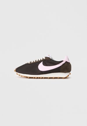 Czarno-brązowy sneaker Nike z jasnoróżowym symbolem Swoosh i detalem na pięcie, białymi sznurówkami oraz gumową podeszwą na jasnoszarym tle.