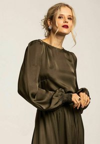 Olivgrön klänning i sidenmaterial med långa puffärmar och smockade ärmar. Har en rund halsringning och en svängd kjol.