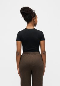 Γυναίκα με σγουρά μαλλιά σε κότσο, που φοράει μαύρο εφαρμοστό μπλουζάκι και καφέ παντελόνι με ψηλή μέση, όρθια με την πλάτη γυρισμένη σε απλό φόντο.