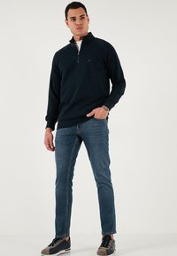 Tengerészkék zipzáras pulóver magas gallérral, sötétkék farmerral és szürke sportcipővel párosítva. Sima anyag és letisztult dizájn.