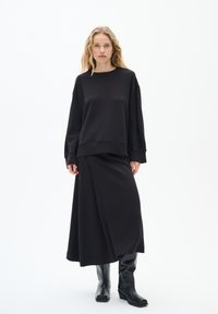 Felpa nera con vestibilità rilassata e scollatura rotonda, abbinata a una lunga gonna nera con design a rouche. Stivali neri fino al ginocchio.
