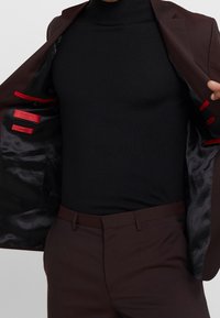 Bruine blazer open om een zwarte turtleneck te onthullen, met rode accenten aan de binnenkant van de blazer. De broek is ook bruin, wat een samenhangende uitstraling creëert.