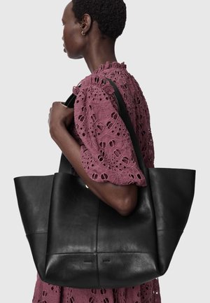 Femme portant une robe ajourée mauve détaillée avec un grand sac cabas en cuir noir sur l'épaule, sur un fond uni.