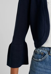 Cardigan cropped bleu marine avec tricot texturé et manches évasées, porté sur un top clair, assorti à un short en denim déchiré.
