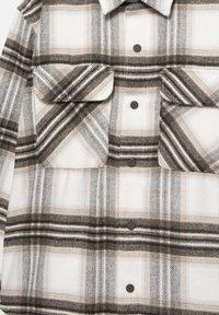 Camicia in flanella a quadri nei toni beige, bianco e nero, con due tasche sul petto e colletto arrotondato, con bottoni scuri sul davanti.
