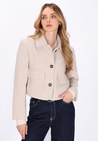 Beige cropped jakke lavet af blødt stof, med krave, to store forlommer og tre knapper, båret over en lys trøje.