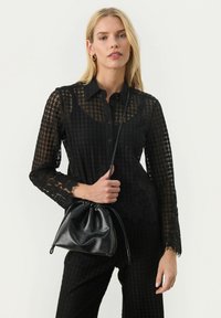 Blusa nera in pizzo con colletto e chiusura a bottoni, caratterizzata da maniche lunghe trasparenti e un motivo tessurizzato. Abbinata a una borsa nera testurizzata.