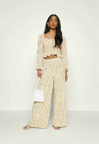 Conjunto coordenado com padrão floral em tecido amarelo claro, apresentando uma blusa cropped de ombros à mostra com Babados e calças de perna larga, acessorizado com uma bolsa branca.