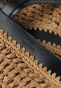 Črni usnjeni trak z vbočenim logotipom na tkanem rjavem materialu z vzorcem teksture in obliko s valovitim robom.