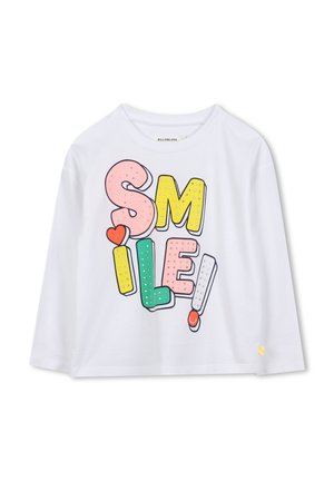 Hvid skjorte med lange ærmer med farverige, store blokbogstaver, der staver "SMIL!", udsmykket med små skinnende nitter og en rød hjerte-detalje på "I'et".