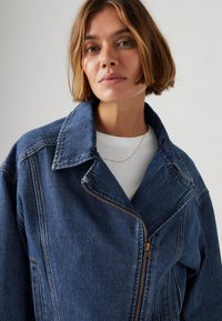 Denim-takki, jossa on suuri kauluksenpää, kultainen vetoketju ja hieman ylisuurta istuvuutta. Ominaisuuksiin kuuluu sininen kangas, jossa on hienovaraisia tekstuurimuutoksia.