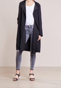 Abrigo negro largo con cuello afilado, combinado con una camiseta blanca sin mangas, jeans ajustados grises y sandalias negras de tacón con acentos de tachuelas.