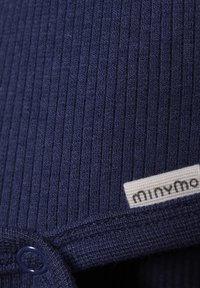 Mørkeblå ribbet stof med en struktureret finish. En rund knapdetalje og et lille hvidt mærke med "minymo" er synlige.
