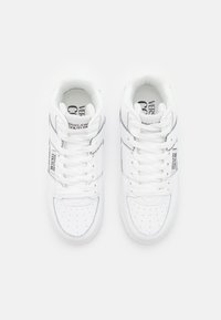 White sneakers with a perforated front, flat laces, and contrasting black branding. Smooth leather exterior with a low-top design.