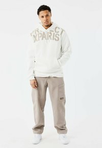 Sudadera con capucha color crema con el texto "PROTECT XPARS" en marrón claro. Combinada con pantalones cargo beige claros y zapatillas blancas.