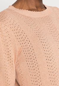 Un pull rose clair avec un motif texturé en maille ouverte et un col côtelé. Le tissu semble doux avec un éclat subtil.
