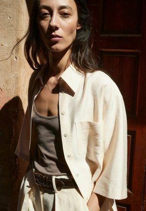 Kobieta o długich ciemnych włosach, ubrana w beżową rozpiętą koszulę na szarym topie i białych spodniach, stoi oparta o teksturowaną ścianę i drewniane drzwi.