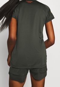 Ciemnozielona sportowa koszulka z krótkimi rękawami, z teksturowanej tkaniny, z pomarańczowym logo "Manchester United" na plecach. Spodenki poniżej.