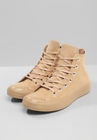 Béžové vysoké tenisky vyrobené z semiše, s gumovou špičkou, plochými tkaničkami a kovovými očky. Texturovaný povrch s hladkými akcenty.