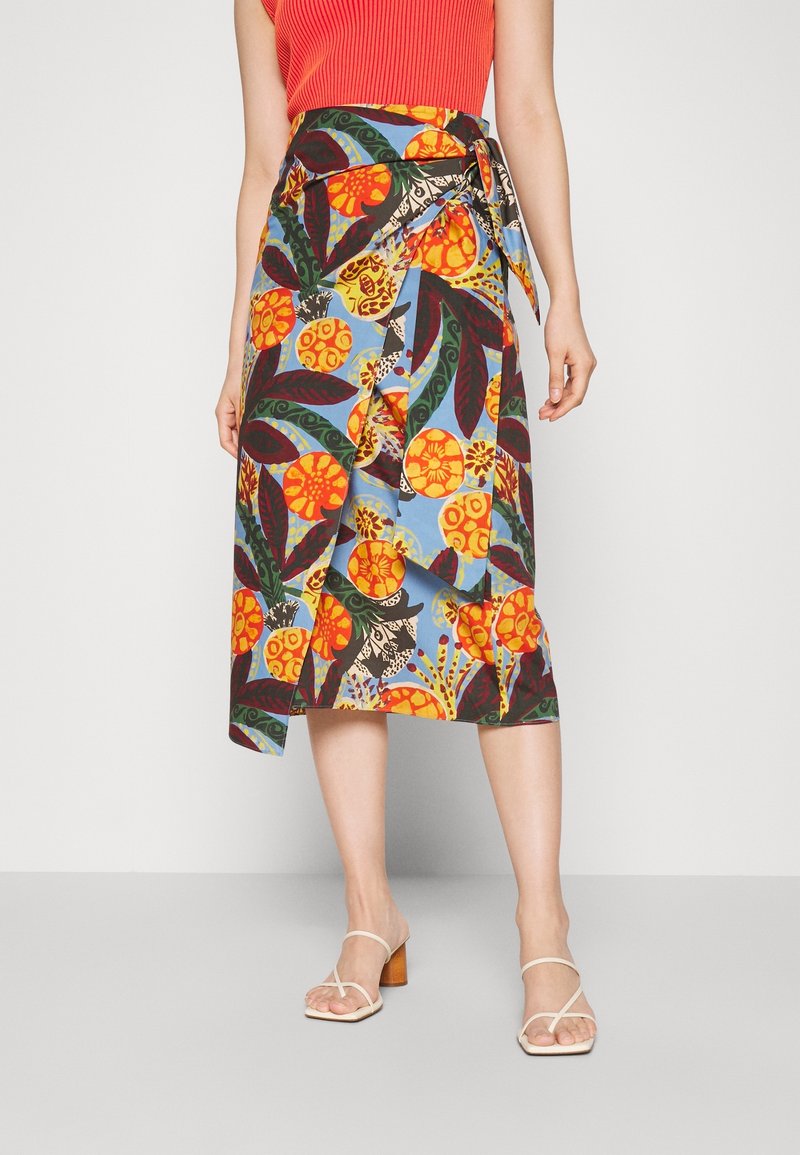 Femme portant une jupe portefeuille avec des motifs floraux orange et verts sur un fond bleu, associée à un haut rouge sans manches et des talons blancs à brides.