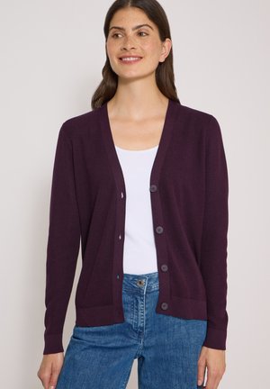 Vrouw die een donkerpaarse cardigan met knopen draagt over een witte top en blauwe spijkerbroek, kijkt iets opzij met een subtiele glimlach.