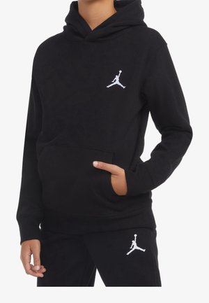 Garçon portant un sweat à capuche noir et un pantalon assorti avec des logos Jumpman blancs, une main dans la poche du sweat à capuche, sur un fond blanc.